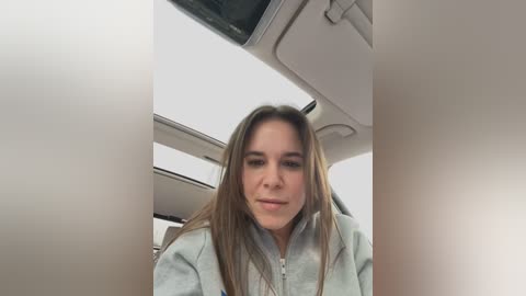 Media: Video of a young woman with long brown hair, wearing a light blue zip-up jacket, seated in a car. The background features a beige interior with a sunroof and light-colored ceiling.