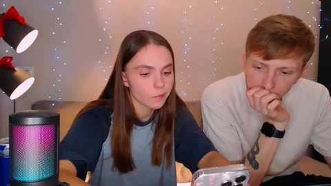 Media: Video of a young woman with long hair in a blue shirt, and a young man with short hair in a white shirt, both looking contemplative at a desk, surrounded by a minimalist, softly lit room.