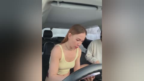 Media: A video of a young woman with light skin and straight, shoulder-length brown hair, wearing a yellow tank top, concentrating on driving a car with a beige interior.