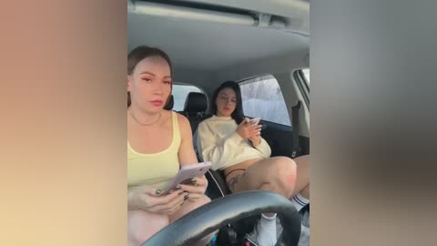 Media: Video of two young women in a car; one with fair skin, light makeup, and a yellow tank top, holding a phone; the other with darker skin, long black hair, and a white sweater, also on a phone.