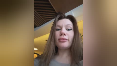 Media: Video of a young Caucasian woman with straight brown hair, light skin, and a neutral expression. She wears a light gray shirt, standing in a warmly lit room with wooden ceiling beams.