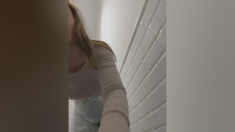 Media: A blurry video of a woman with light skin and blonde hair, wearing a beige top and blue apron, cleaning white-tiled walls in a dimly lit bathroom.