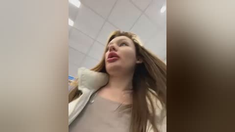 Media: Video of a woman with long, straight brown hair, wearing a beige top, sticking her tongue out. Background shows a white ceiling with square tiles. Image is blurry and out of focus.