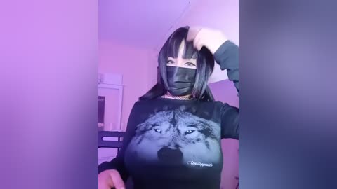 Media: Video of a pale-skinned woman with straight black hair, wearing a black face mask and a black shirt featuring a large wolf's head graphic. She stands indoors under purple lighting, with a hand in her hair.