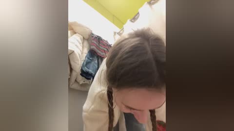 Media: Video of a young woman with long brown hair and a tan jacket, bent over in a small, dimly-lit room with a green ceiling and clothes hanging on the wall.