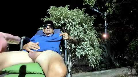 Media: A video of a middle-aged, light-skinned man in a blue polo and sunglasses, sitting in a green chair outdoors at night, with a tree and hanging lanterns in the background.