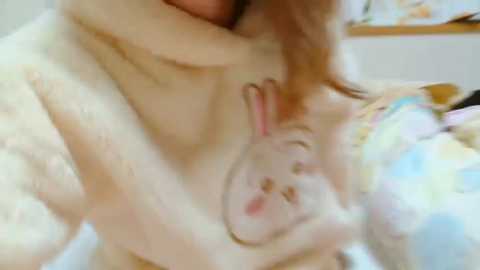 Media: Video of a young woman with light skin and short brown hair, lying on a bed with a fluffy white dog cuddling her head. She is wearing a light-colored top, and the background is blurred.