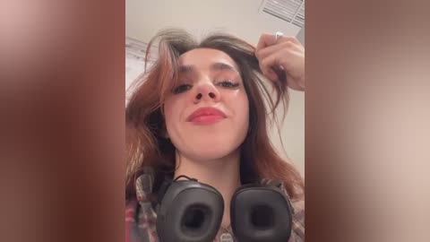 Media: A video of a young woman with fair skin, brown hair, and pink lipstick, wearing headphones, taken from a close-up angle. She appears relaxed, with a slight smile, in a plain room with a vent and white wall.