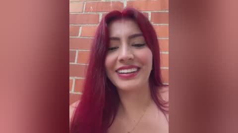 Media: A video of a smiling woman with long, straight, deep red hair, fair skin, and red lipstick, set against a brick wall. The background is blurred, focusing attention on her happy expression.