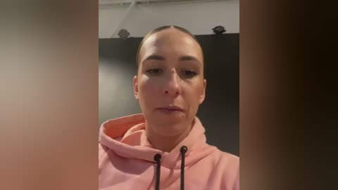 Media: Video of a woman with light skin, high cheekbones, and a slim figure, wearing a pink hoodie, standing in a dimly lit room with black curtains.