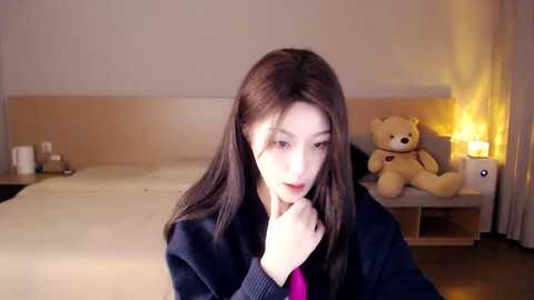 Media: Video of a young Asian woman with long dark hair, wearing a black sweater, standing in a cozy, dimly lit bedroom with a white bed, beige wall, teddy bear, and nightstand lamp.