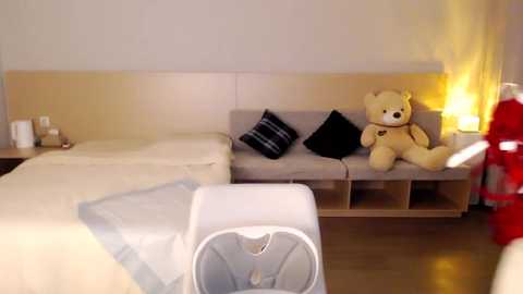 Media: A video of a modern, minimalist bedroom featuring a beige headboard, a white bed with a gray blanket, two black pillows, a teddy bear, and a yellow lamp.