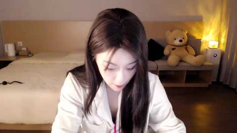 Media: Video of a young Asian woman with long black hair, wearing a white blouse, sitting on a bed in a dimly-lit bedroom with a teddy bear, a nightstand, and a white bedspread.