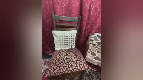 Media: Video of an old wooden chair with a white crochet cushion and maroon patterned seat, set against red floral-patterned curtains. The background includes a partially visible second chair with a similar pattern.