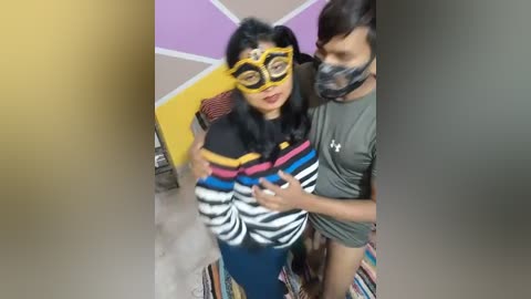 A video showing two people in a room with colorful, abstract walls. One wears a yellow mask, black shirt, and striped leggings, while the other wears a green shirt.