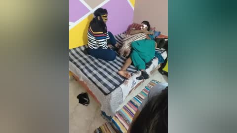 Video of a woman in a striped shirt and dark pants sitting on a bed with a black and white striped blanket, while a man in green pajamas lies on his stomach beside her. The room has purple and yellow walls with a white geometric pattern.