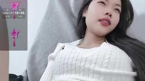 Media: Video of an Asian woman with long black hair, fair skin, wearing a white ribbed sweater, leaning back, in a soft-focus, indoor setting.