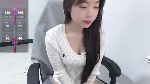 Media: Video of an East Asian woman with long black hair, fair skin, and a slender physique, wearing a white ribbed sweater, sitting on a black office chair in a plain white room.