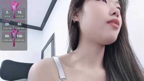 Media: Video of an Asian woman with long, straight brown hair, light skin, and wearing a white tank top, looking pensive. Background shows a medical chart and a chair.