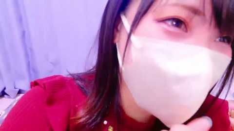 Media: Video of a young woman with long dark hair wearing a red shirt and a white mask, looking to the side with a neutral expression, in a blurry, light-colored background.