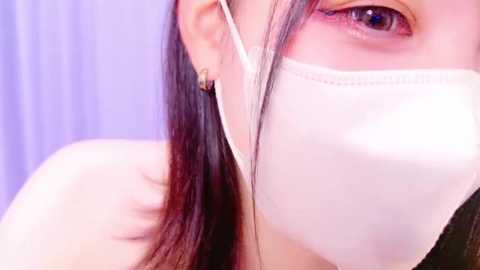 Media: Video of a person with light skin, wearing a white face mask, revealing one eye with a piercing blue iris. They have long, straight, dark hair, and are wearing a simple black outfit. The background is a soft, pastel purple.