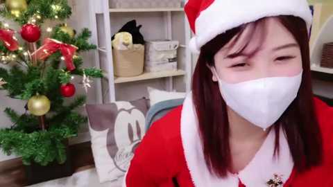 Media: Video of an Asian woman with straight, dark hair, wearing a red Santa hat and a white mask, standing in a festive room with a decorated Christmas tree and shelves filled with holiday decor.