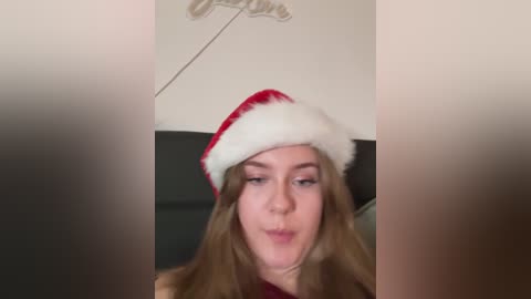 Media: A video of a young woman with fair skin and long brown hair, wearing a red Santa hat, making a kissy face. The background shows a dark leather couch and a beige wall with a faintly visible decorative piece.