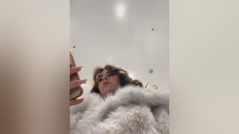 Media: Video of a young woman with light skin and wavy brown hair, wearing a fluffy white coat, holding a smartphone, gazing upward in a minimalist, bright room.