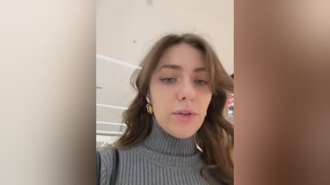 Media: Video of a young Caucasian woman with wavy brown hair, wearing a gray ribbed turtleneck sweater, standing in a bathroom with white walls and a shower curtain.