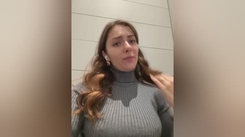 Media: Video of a young Caucasian woman with wavy brown hair, wearing a grey ribbed turtleneck sweater, standing against a beige tiled wall, looking slightly to the side.