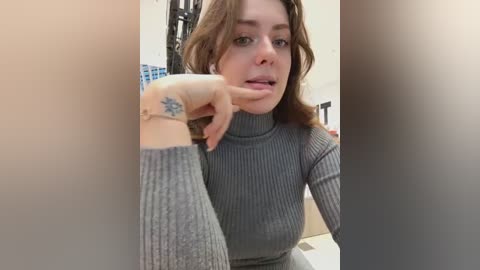 Media: A video of a young Caucasian woman with shoulder-length brown hair, wearing a gray ribbed turtleneck sweater, holding her finger to her lips. She has a tattoo on her wrist. The background is blurred.