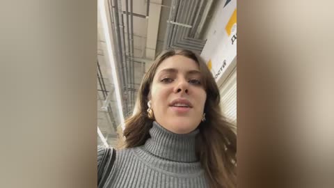 Media: A video of a young Caucasian woman with long brown hair, wearing a grey ribbed turtleneck sweater, standing in a dimly lit, industrial corridor with exposed pipes and yellow and white wall signs.