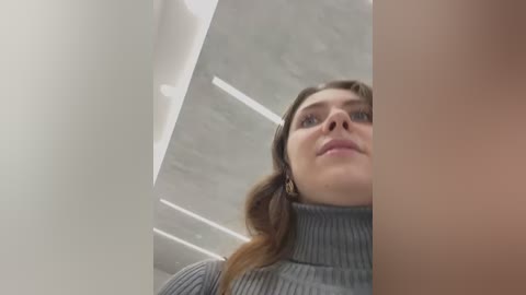 Media: Video of a young Caucasian woman with long brown hair, wearing a grey ribbed turtleneck sweater, looking up at a modern white ceiling with recessed lighting. The image is angled from below, emphasizing her face and upper body.