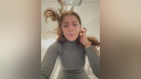 Media: Video of a young woman with fair skin, long wavy brown hair, wearing a gray ribbed turtleneck sweater, standing in a dimly lit room with beige walls, her hair flying in front of her face.