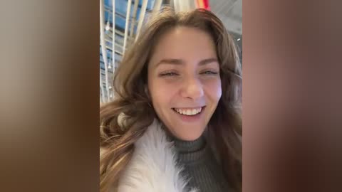 Media: A video of a young woman with light skin, long brown hair, and a smile, wearing a grey turtleneck sweater, standing in a modern, indoor space with metal railings and a blue background.