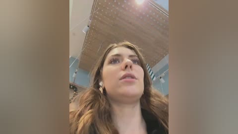 Media: A video of a young woman with fair skin, long brown hair, and wearing a black top, looking upwards with a serene expression. The background features a wooden ceiling with a skylight and a white wall.