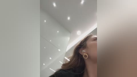 Media: Video of a woman with light skin and brown hair wearing a black dress, standing in a modern, brightly lit room with white walls and recessed lighting.