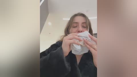 Media: A video of a young woman with light skin and long brown hair, wearing a black robe, blowing her nose into a white tissue. The background is a plain, light-colored room with minimal decor.