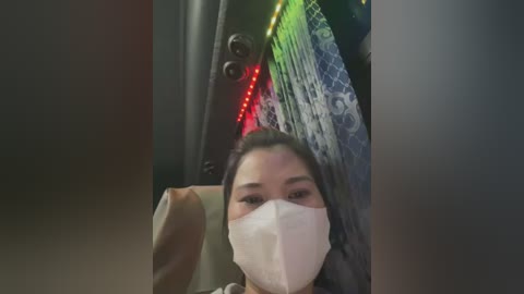 Media: Video of a woman with long dark hair, wearing a white mask, sitting in a car. The background shows a colorful, patterned fabric hanging behind her, with a neon sign and red lights.