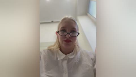 Media: Video of a blonde woman with fair skin, wearing glasses, red lipstick, and a white shirt, looking through a narrow gap between two white walls in a bright room with a window.