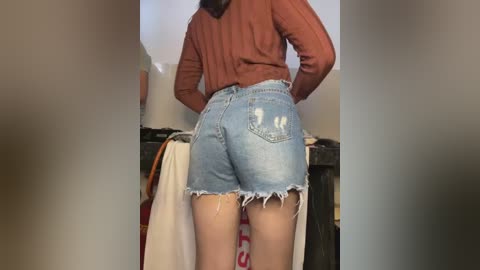 Media: A video of a woman with medium skin tone and long brown hair, wearing a brown sweater and ripped light blue denim shorts, standing in a dimly lit room with a towel and dark wooden shelf in the background.