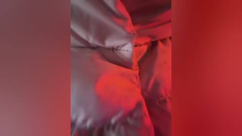 Media: Video of a close-up, tightly cropped section of a person's body, covered in a shiny, metallic silver fabric, illuminated by intense red lighting. The background is blurred, focusing on the reflective surface and the dramatic color contrast.