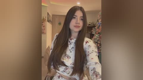 Media: Video of a young woman with long, straight brown hair, wearing a floral-patterned dress, sitting in a cozy, warmly lit living room with Christmas decorations.