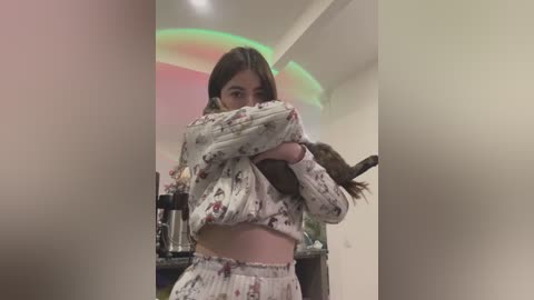 Media: Video of a young woman with long brown hair, wearing a floral pajama set, hugging a small black cat in a modern, dimly lit room with a soft, greenish light.