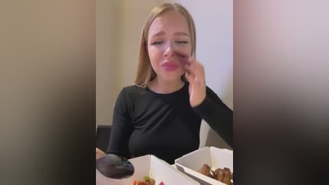 Video of a blonde woman with fair skin, wearing a black long-sleeved top, wiping a tear with her left hand while holding a black olive in her right. She sits at a table with a white box of fried chicken.