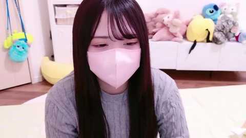 Media: Video of an East Asian woman with long dark hair, wearing a white face mask, light grey sweater, in a plush toy-filled, pastel-colored bedroom.