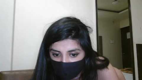 Media: A video of a woman with long black hair, wearing a black mask covering her mouth, standing indoors. She has brown eyes and fair skin. Background shows a white wall, a door, and a mirror.