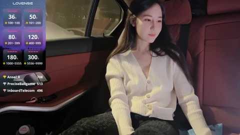 Media: Video of a young woman with long black hair, fair skin, wearing a beige cardigan and black pants, sitting in a red car interior, with a smartphone screen displaying her social media profile.