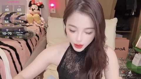 Media: Video of a fair-skinned, slender woman with long brown hair and red lipstick, wearing a black lace top, sitting on a bed with a Minnie Mouse pillow. Background shows a cluttered room with a pink lamp and clothing.