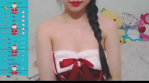 Media: A young Asian woman with long braided hair, fair skin, and red lipstick wears a strapless red and white Santa-themed top. Background shows a blue wall with gaming interface and plush toys.